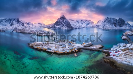 Aerial view of blue sea, snowy mountains, rocks, village, buildings, rorbu, road, bridge, colorful cloudy sky at sunset in winter.  Hamnoy in Lofoten islands, Norway. Panoramic landscape. Top view