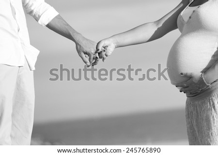 Black and white photo of a pregnant woman and her husband holding their hands at the beach