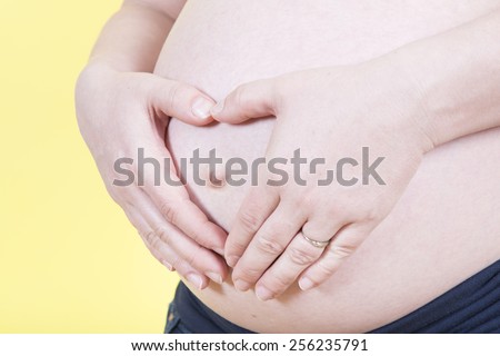 Pregnant woman with her hands on her belly and her fingers are heart shaped in front of a yellow background