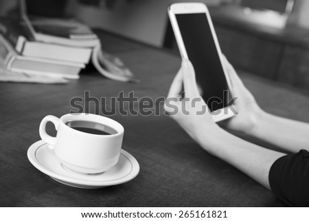 Black and white photo of a woman holding a tablet with a no name coffee cup and a stack of books in background on a wood table with shallow depth of field