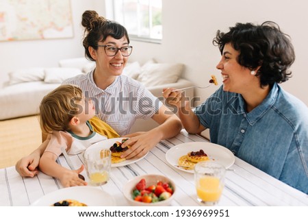 Female couple with son having breakfast at home. Smiling women looking at each other while eating breakfast with their son watching.