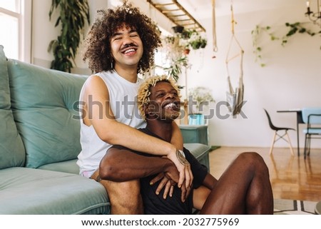 Queer couple embracing each other indoors. Two romantic young male lovers smiling cheerfully while sitting together in their living room. Young gay couple bonding at home.