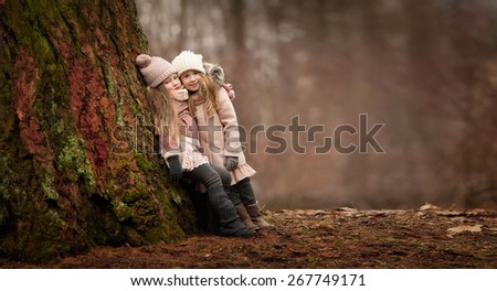 two little girls friends with long blond hair in coats and hats are standing arms in arms and hugging near the big tree in the forest