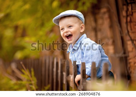 portrait of little laughing boy in the blue shirt and  cap is standing near the wooden fence in the nature