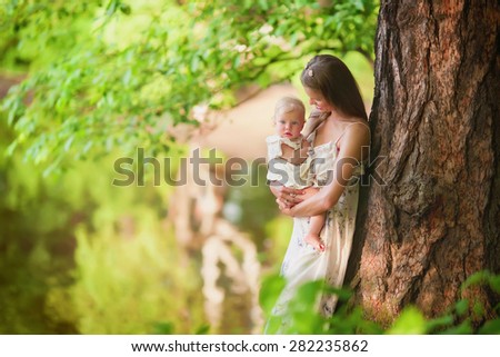 beautiful smiling mother is standing under the tree and holds her little baby girl daughter in the park at summer time