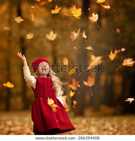 little laughing pretty girl with long blond hair in red hat and red dress throws yellow leaves in autumn park