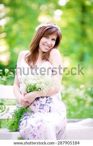 Beautiful smiling pregnant woman with long hair sitting in the park with white flowers in her hands at summer time