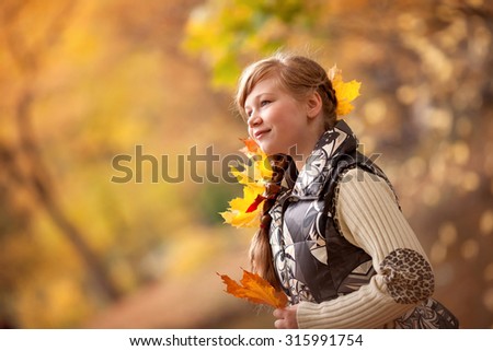 little child girl in sweater and vest standing in the autumn park and looking with leaves on her hands