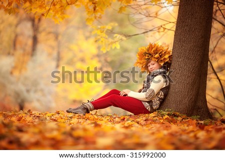 little child girl in sweater and vest sitting in the beautiful autumn park near the tree with wreath of leaves on her head
