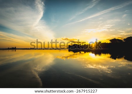 Sun rise from the trees and reflect together with clouds and sky in the water surface. Striking sunset above the ocean bay with bright clouds and sky.