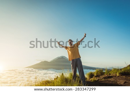 Man stay on the top of hill and rise his hands up with white clouds and peak of volcano on background