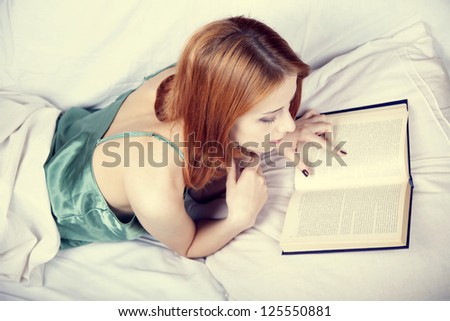 Pretty red-haired woman in lying in the bed and reading book. Studio shot.