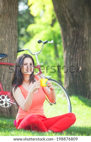 Brunette girl with cup of coffee in the park and with bicycle on background.