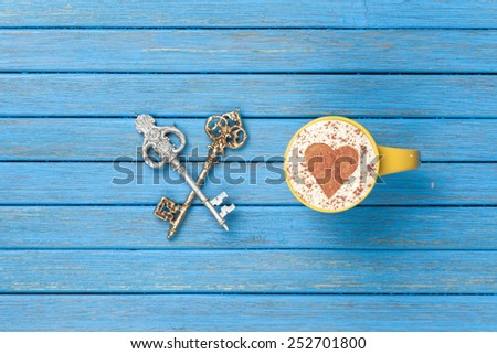 Cup of Cappuccino with heart shape symbol and two keys on blue wooden background.