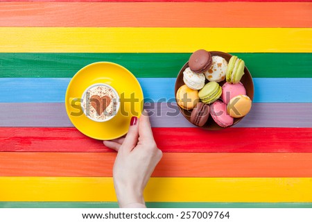 Female hands holding cup of coffee with heart shape near macarons on color background.