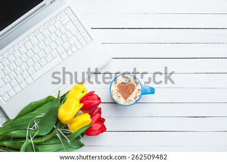 Cup of cappuccino with heart shape and computer, tulips on wooden background.