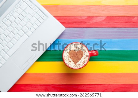 Cup of cappuccino with heart shape and computer on multicolor wooden background.