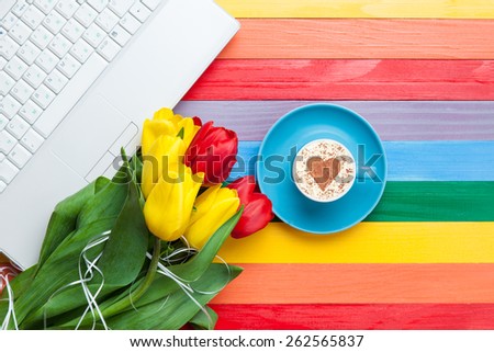 Cup of cappuccino with heart shape and computer, tulips on multicolor wooden background.