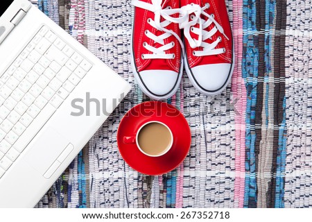 Cup of cappuccino near red gumshoes and computer on carpet background. Above view