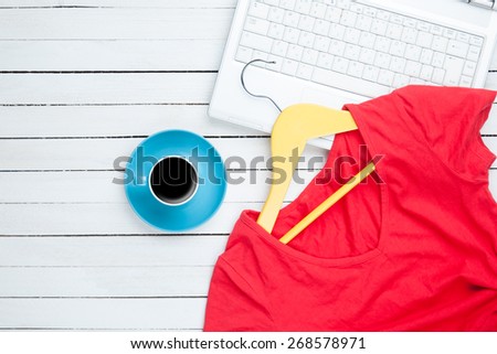 Cup of coffee and hanger with red dress near computer on white wooden table.