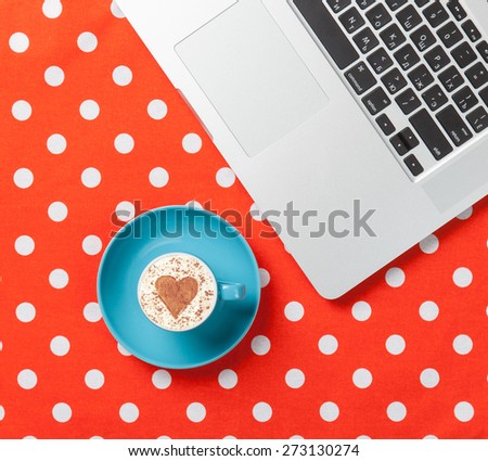 Cup of cappuccino with heart shape and laptop on red polka dot background.