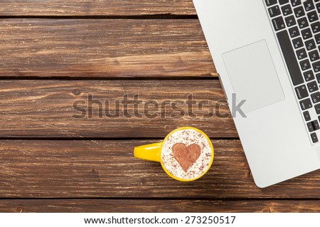 Cup of cappuccino with heart shape and laptop on wooden table.