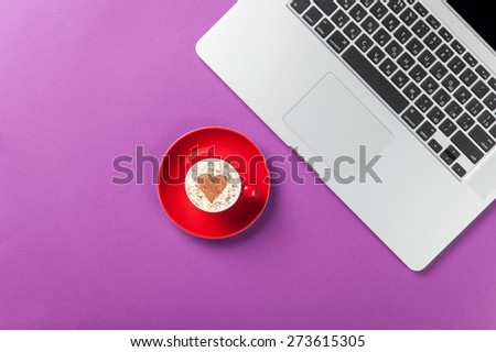 Cup of cappuccino with heart shape and laptop on violet background.