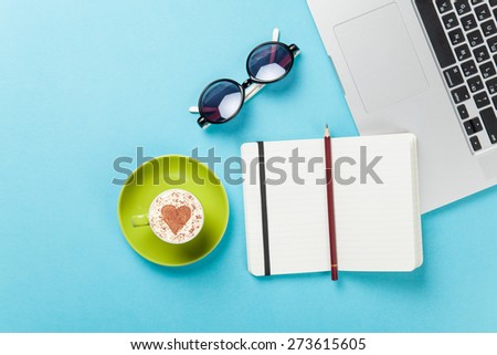 Cup of cappuccino with heart shape and laptop near glasses on blue background.