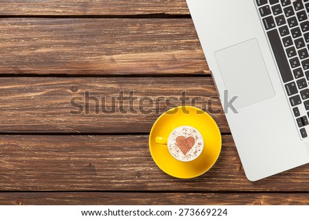 Cup of cappuccino with heart shape and laptop on wooden table.