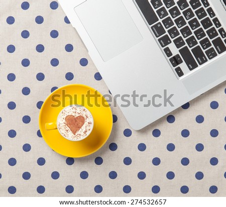 Cup of cappuccino with heart shape and laptop on polka dot background.