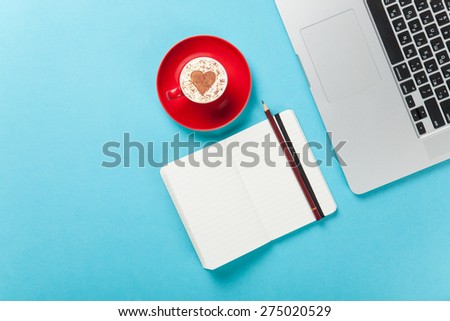 Cup of cappuccino with heart shape and laptop with note on blue background.