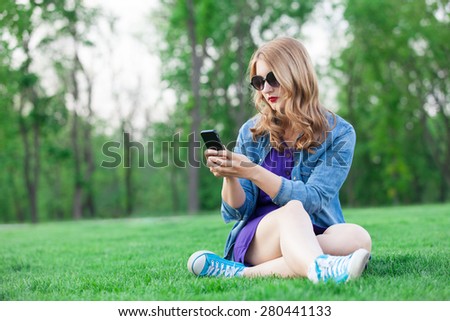 Beautiful blonde women sitting on green grass and speaking by the phone in summer time in the park.