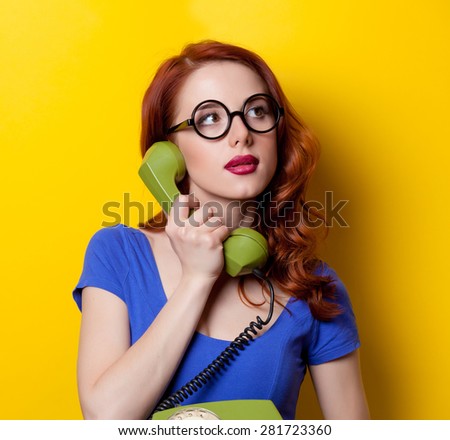 Young redhead girl in blue dress with dial phone on yellow background.