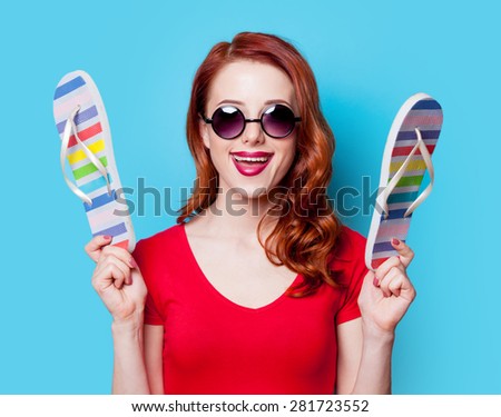 Happy redhead girl in sunglasses with flip flops and red dress on blue background