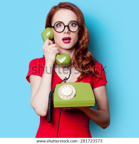 Smiling redhead girl in red dress with green dial phone on blue background.