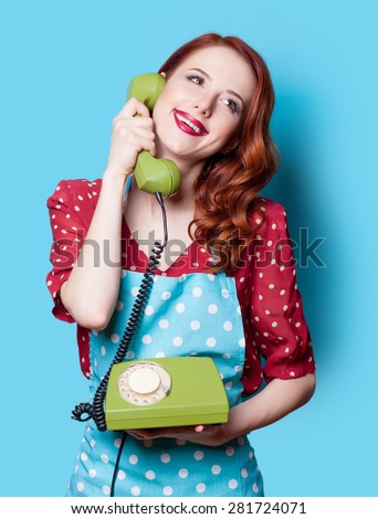 Smiling redhead girl in red polka dot dress with green dial phone on blue background.