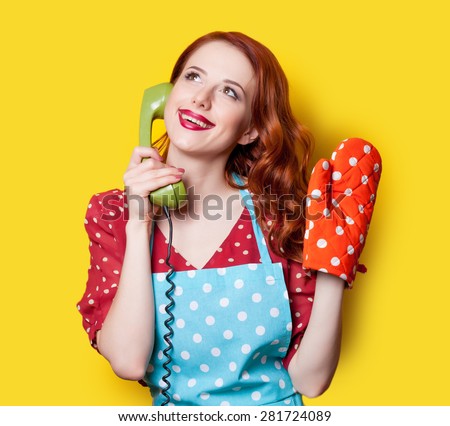 Smiling redhead girl in red polka dot dress and blue apron with green dial phone on yellow background.