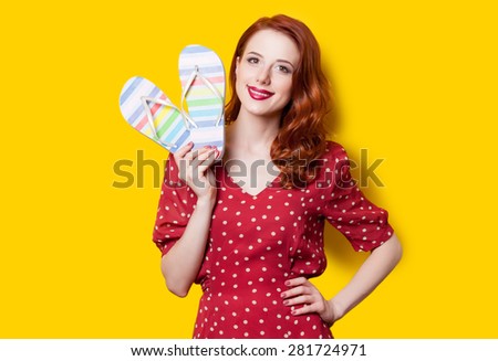 Smiling redhead girl in red polka dot dress with flip flops on yellow background.