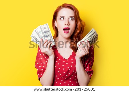 Surprised redhead girl in red polka dot dress with money on yellow background.