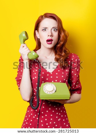 Surprised redhead girl in red polka dot dress with green dial phone on yellow background.