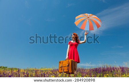 Portrait of a beautiful redhead girl in red dress with umbrella and suitcase on the meadow.