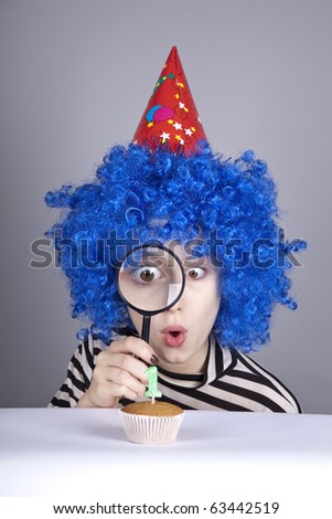 Funny girl with blue hair and striped knitted jacket looking through loupe at cake for birthday with one candle.