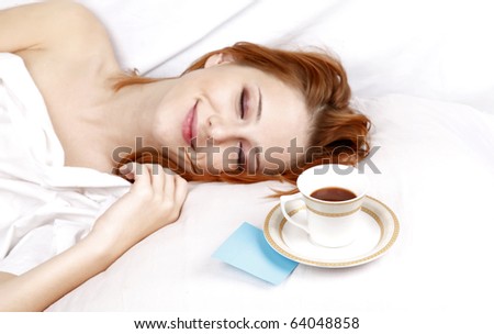 Pretty red-haired sleeping woman in white nightie lying in the bed near cup of coffee.