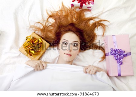 Surprised red-haired girl in bed with gifts. Studio shot.