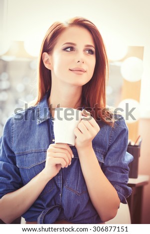 Young redhead women with white cup of coffee sitting near the mirror in dressing room