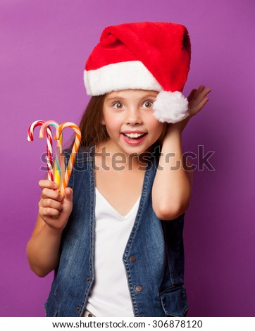 Beautiful young girl in red Santas hat with candy on violet background.