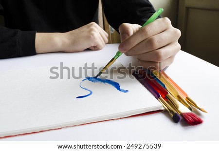 The left hand draws a brush with blue paint on paper in an album with several colorful brushes near it.