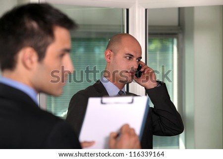 Businessman holding a clip-board whilst colleaguemake a phonre dall