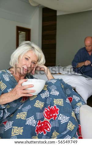 Elderly woman in bathrobe sitting in an armchair having breakfast in front of a man sitting on a bed while reading a magazine