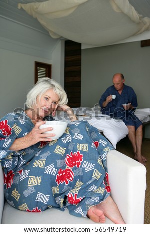 Elderly woman in bathrobe sitting in a chair having breakfast in front of a man sitting on a bed while reading a magazine